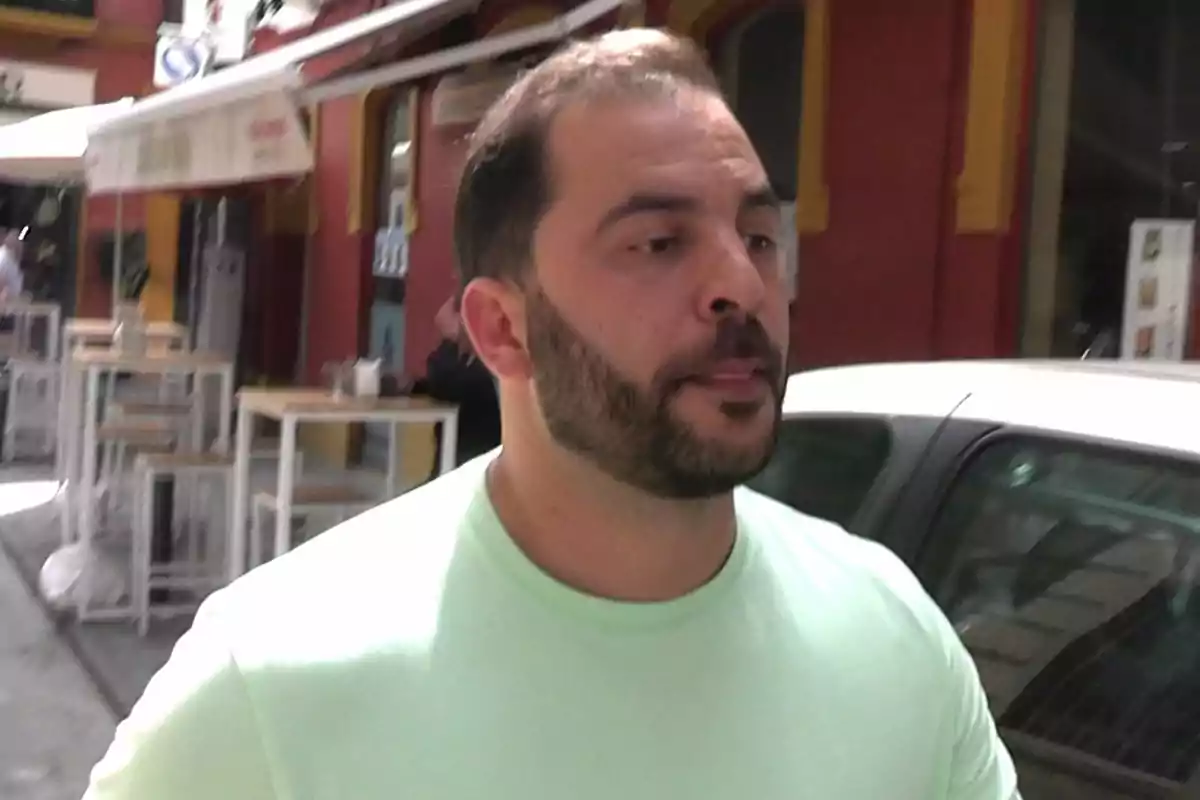 Un hombre con barba y camiseta clara caminando por una calle con mesas al aire libre y edificios coloridos. Un hombre con barba y camiseta clara caminando por una calle con mesas al aire libre y edificios coloridos.