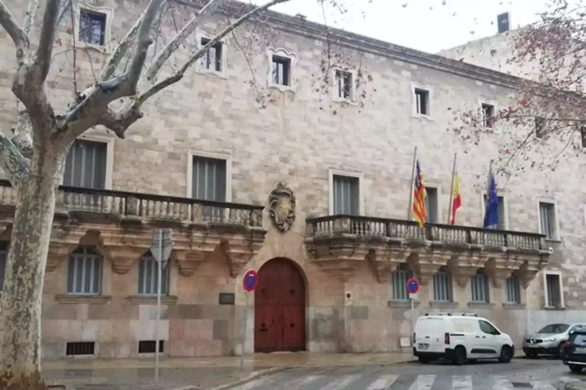 Fachada de un edificio histórico de piedra con balcones, varias banderas y árboles sin hojas en la acera Fachada de un edificio histórico de piedra con balcones, varias banderas y árboles sin hojas en la acera