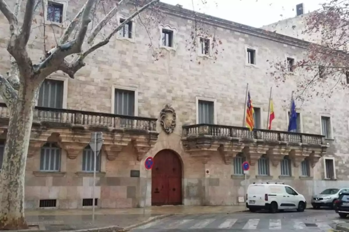 Fachada de un edificio histórico de piedra con balcones, varias banderas y una puerta de madera roja, en una calle con árboles y vehículos estacionados.