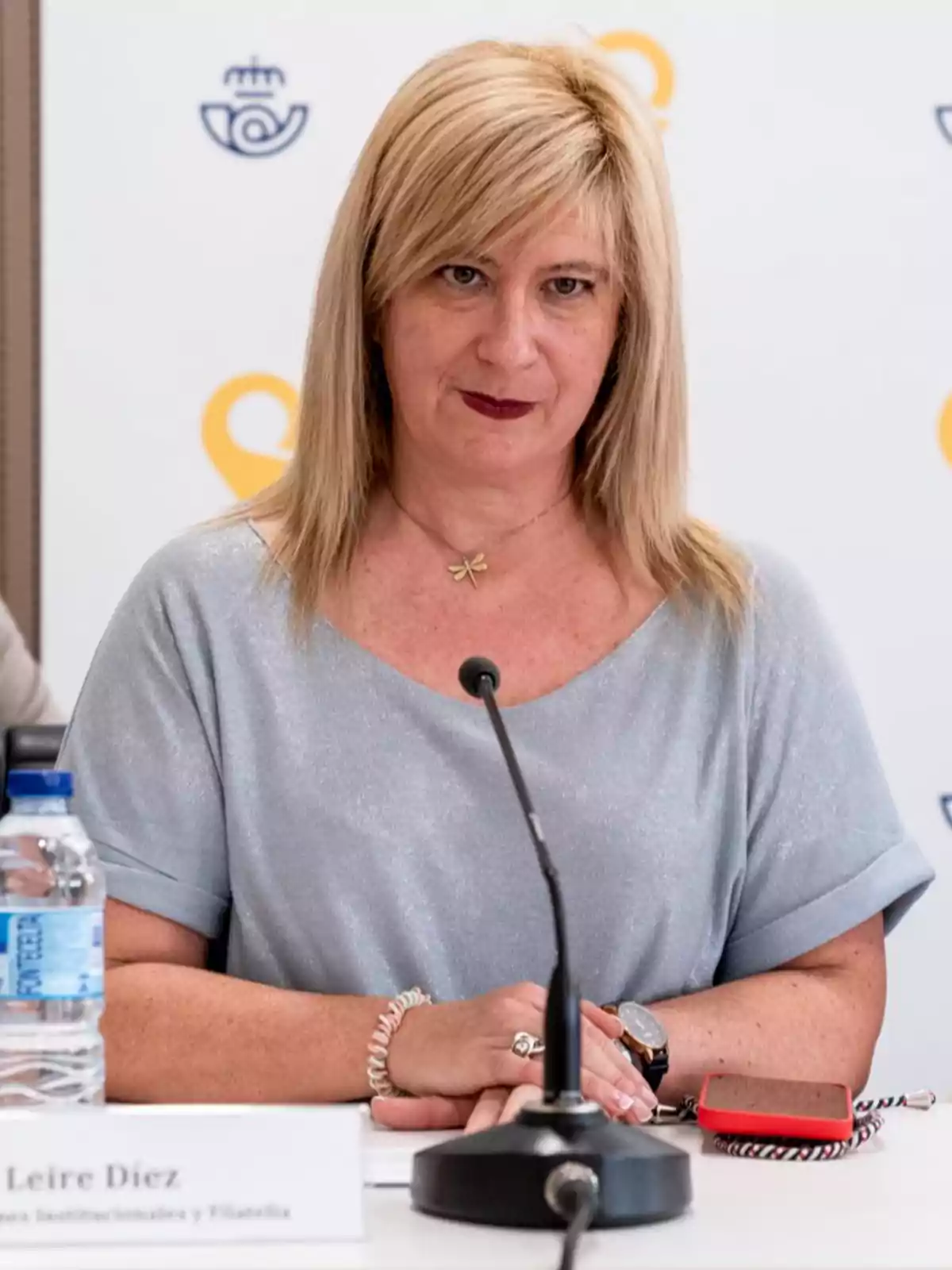 Mujer rubia sentada en una mesa durante una conferencia, con un micrófono frente a ella y una botella de agua a su lado Mujer rubia sentada en una mesa durante una conferencia, con un micrófono frente a ella y una botella de agua a su lado