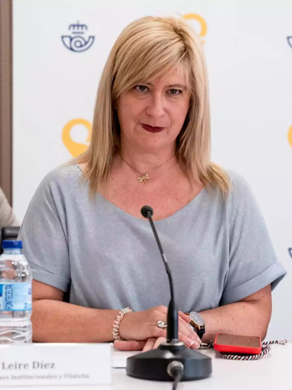 Mujer rubia sentada en una mesa durante una conferencia con un micrófono frente a ella y una botella de agua a su lado Mujer rubia sentada en una mesa durante una conferencia con un micrófono frente a ella y una botella de agua a su lado