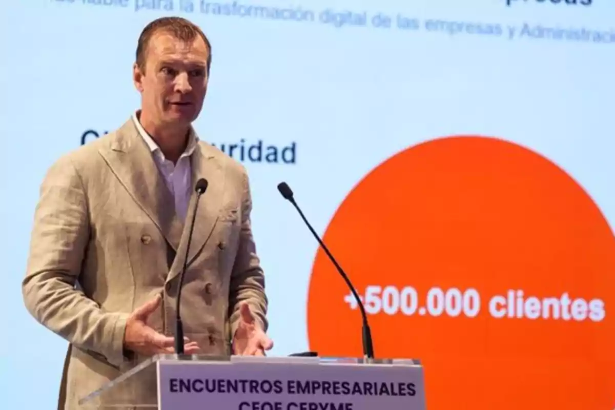 Hombre de traje beige hablando en un podio durante un evento empresarial con una pantalla de fondo que muestra un gráfico naranja y el texto más de 500000 clientes Hombre de traje beige hablando en un podio durante un evento empresarial con una pantalla de fondo que muestra un gráfico naranja y el texto más de 500000 clientes