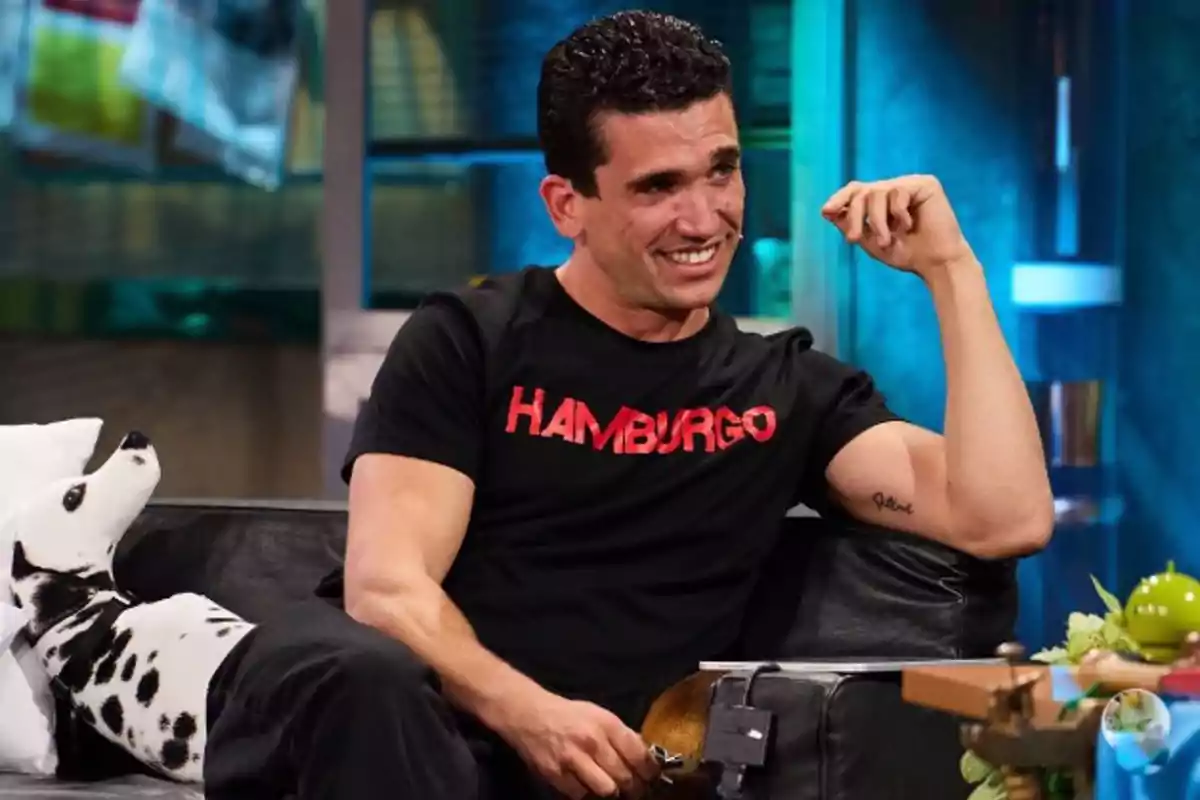Hombre sentado en un sofá negro sonriendo y usando una camiseta negra con la palabra Hamburgo en letras rojas, junto a un cojín con forma de perro dálmata Hombre sentado en un sofá negro sonriendo y usando una camiseta negra con la palabra Hamburgo en letras rojas, junto a un cojín con forma de perro dálmata