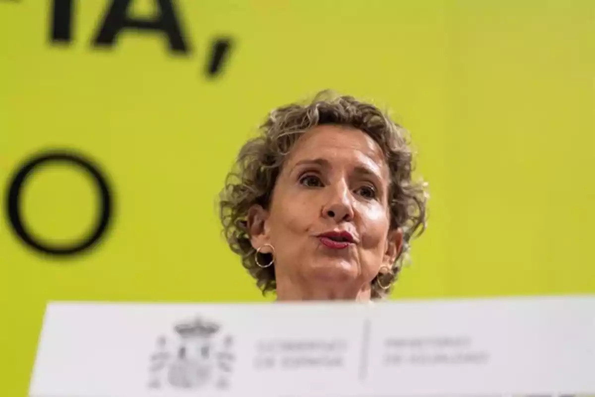 Mujer de cabello corto y rizado hablando en una conferencia con fondo amarillo y un cartel del gobierno de España delante de ella Mujer de cabello corto y rizado hablando en una conferencia con fondo amarillo y un cartel del gobierno de España delante de ella