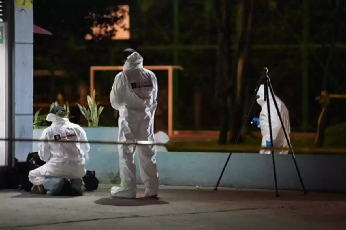 Tres personas con trajes forenses trabajan en una escena acordonada durante la noche Tres personas con trajes forenses trabajan en una escena acordonada durante la noche
