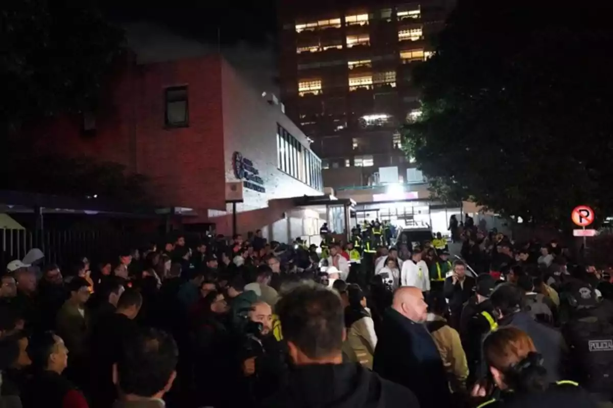 Una multitud de personas y policías se reúne afuera de un edificio durante la noche, con luces encendidas y ambiente de emergencia. Una multitud de personas y policías se reúne afuera de un edificio durante la noche, con luces encendidas y ambiente de emergencia.