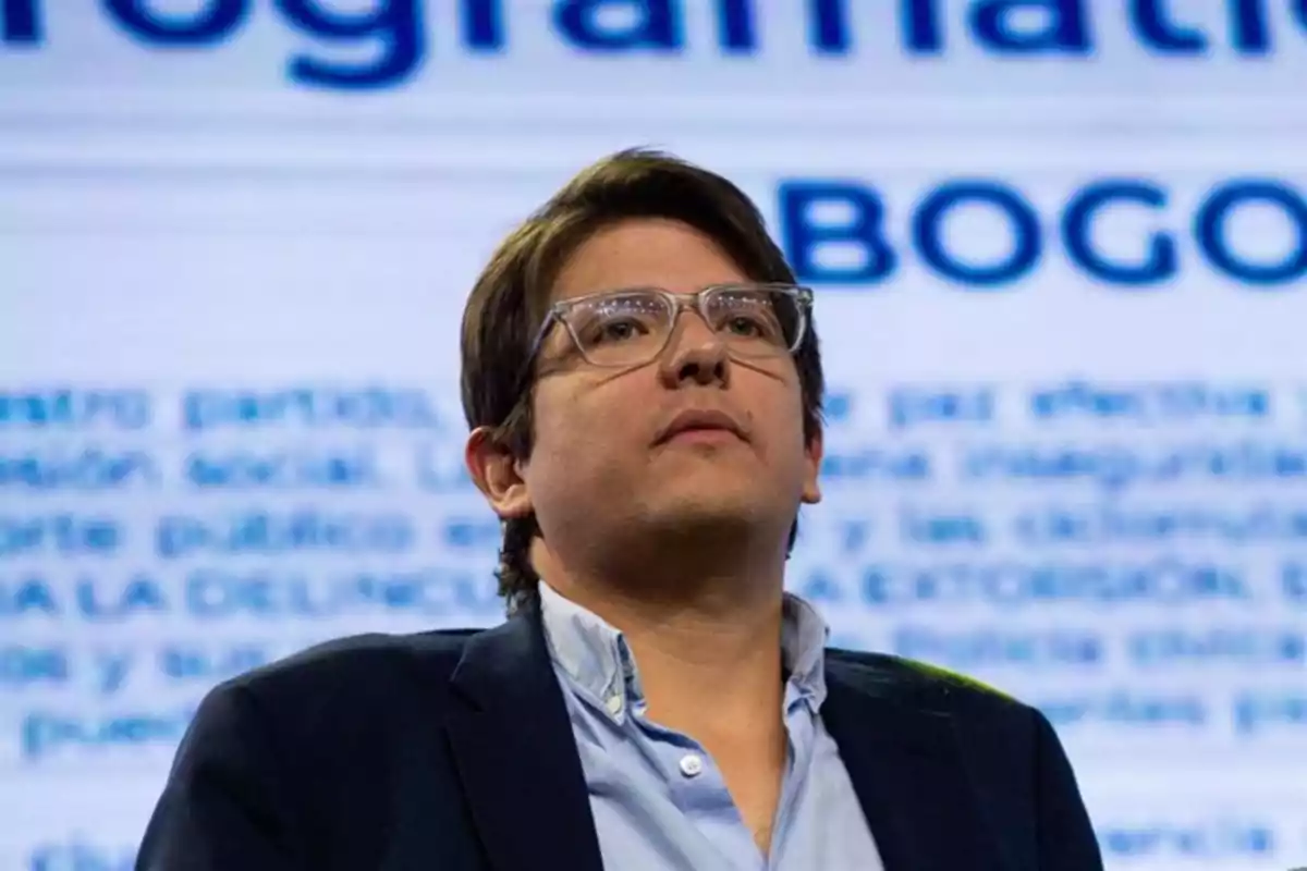 Hombre joven con gafas y saco oscuro mirando hacia arriba con fondo de texto desenfocado donde se lee la palabra Bogotá Hombre joven con gafas y saco oscuro mirando hacia arriba con fondo de texto desenfocado donde se lee la palabra Bogotá