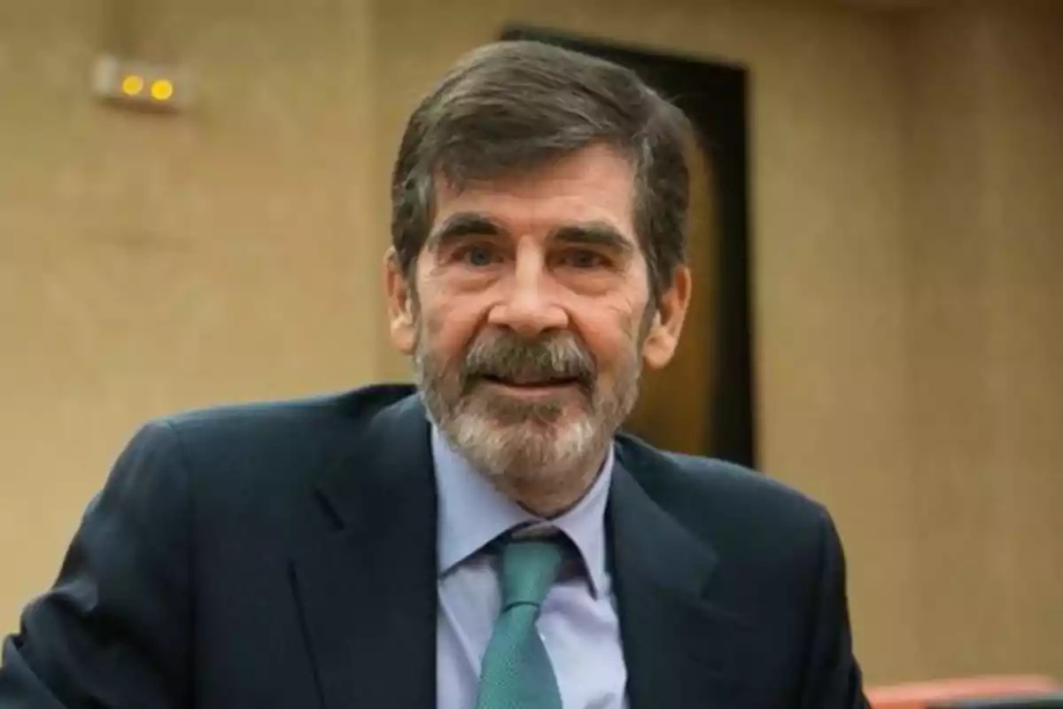 Hombre de mediana edad con barba y cabello canoso vistiendo traje oscuro y corbata verde en un entorno interior