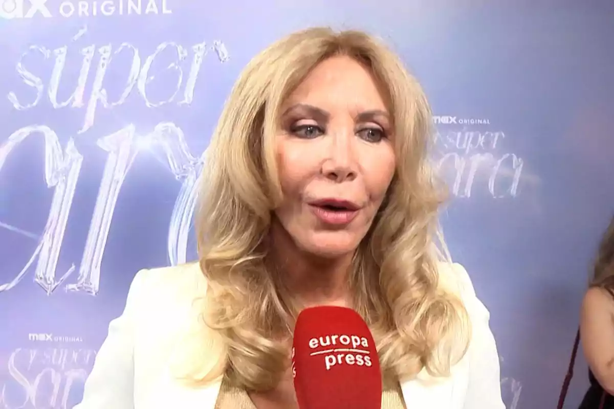 Mujer rubia con cabello ondulado hablando frente a un micrófono rojo de europa press en un evento con fondo azul claro Mujer rubia con cabello ondulado hablando frente a un micrófono rojo de europa press en un evento con fondo azul claro