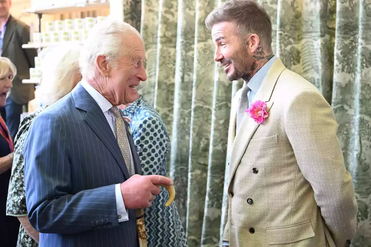 Dos hombres elegantemente vestidos conversan y sonríen en un evento social, uno lleva un traje a rayas y el otro un saco beige con una flor rosa en la solapa Dos hombres elegantemente vestidos conversan y sonríen en un evento social, uno lleva un traje a rayas y el otro un saco beige con una flor rosa en la solapa