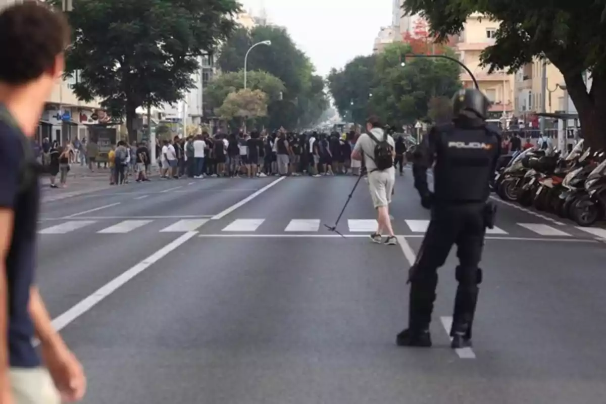 Un grupo de personas se reúne en medio de una calle mientras un policía con equipo antidisturbios observa y un camarógrafo graba la escena Un grupo de personas se reúne en medio de una calle mientras un policía con equipo antidisturbios observa y un camarógrafo graba la escena