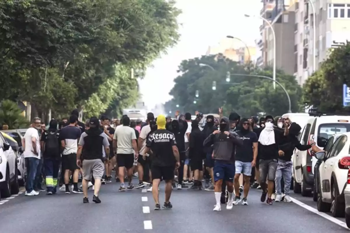 Un grupo numeroso de personas camina por el centro de una calle rodeada de árboles y edificios, algunas llevan el rostro cubierto y visten ropa casual Un grupo numeroso de personas camina por el centro de una calle rodeada de árboles y edificios, algunas llevan el rostro cubierto y visten ropa casual