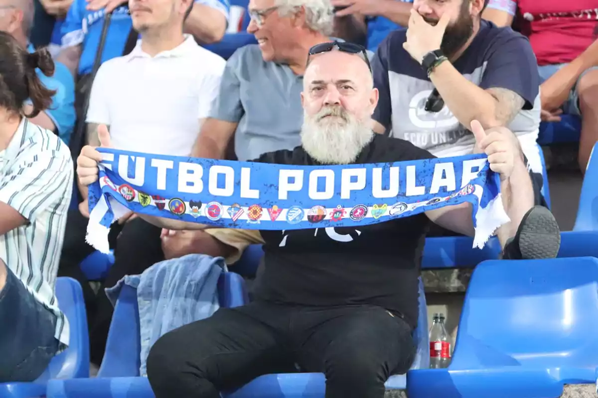 Un hombre con barba blanca sostiene una bufanda azul que dice "FÚTBOL POPULAR" en un estadio lleno de gente.