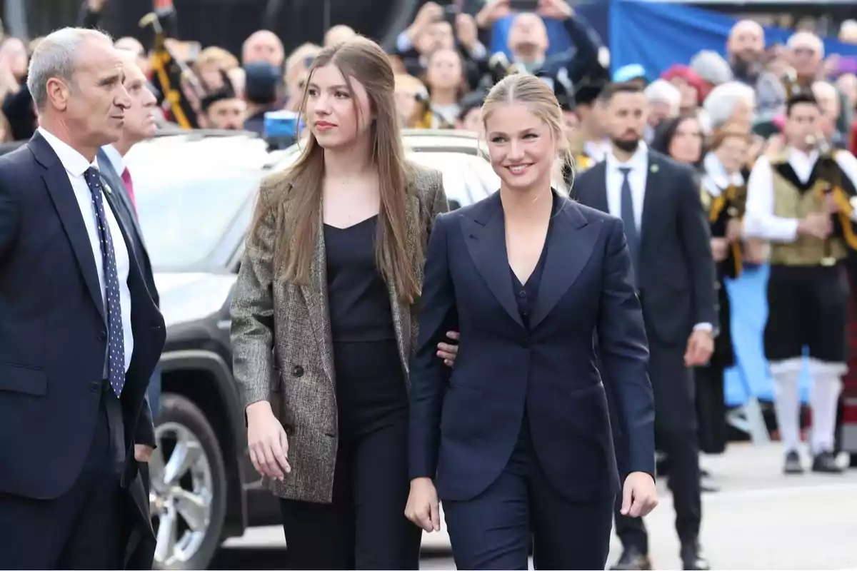 Un grupo de personas bien vestidas camina en un evento al aire libre con una multitud de fondo.