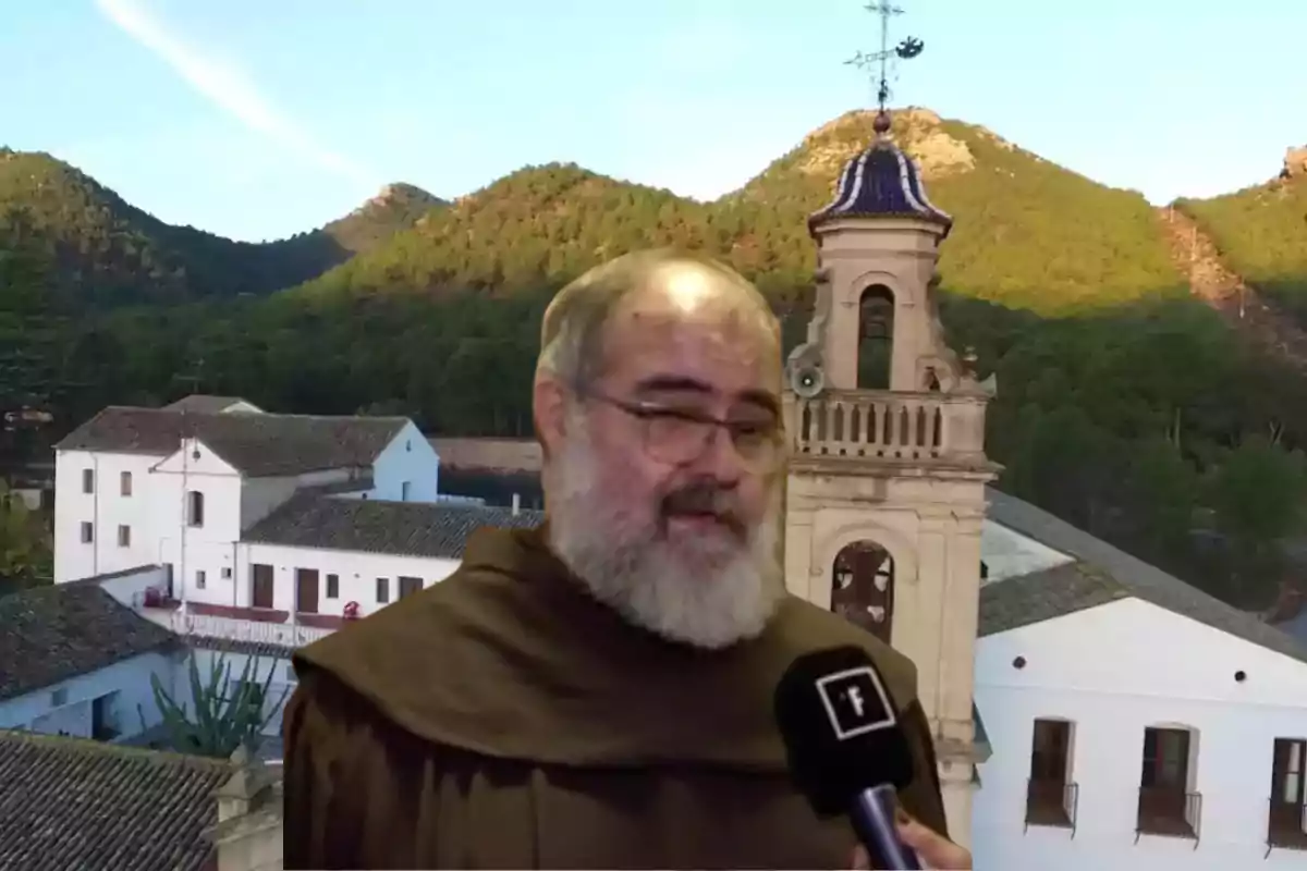 Un hombre con hábito religioso es entrevistado frente a un edificio con campanario y un paisaje montañoso al fondo.