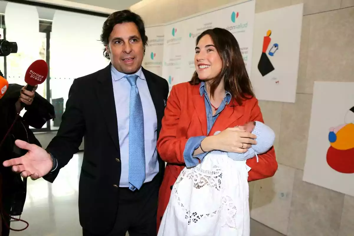 Fran Rivera y Lourdes Montes sonriente sostiene a un bebé envuelto en una manta blanca mientras son entrevistados por la prensa en un hospital. Fran Rivera y Lourdes Montes sonriente sostiene a un bebé envuelto en una manta blanca mientras son entrevistados por la prensa en un hospital.