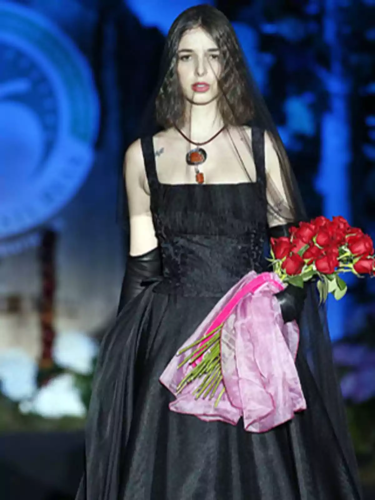 Una modelo desfila con un vestido negro, guantes largos y un collar, sosteniendo un ramo de rosas rojas envueltas en una tela rosa.