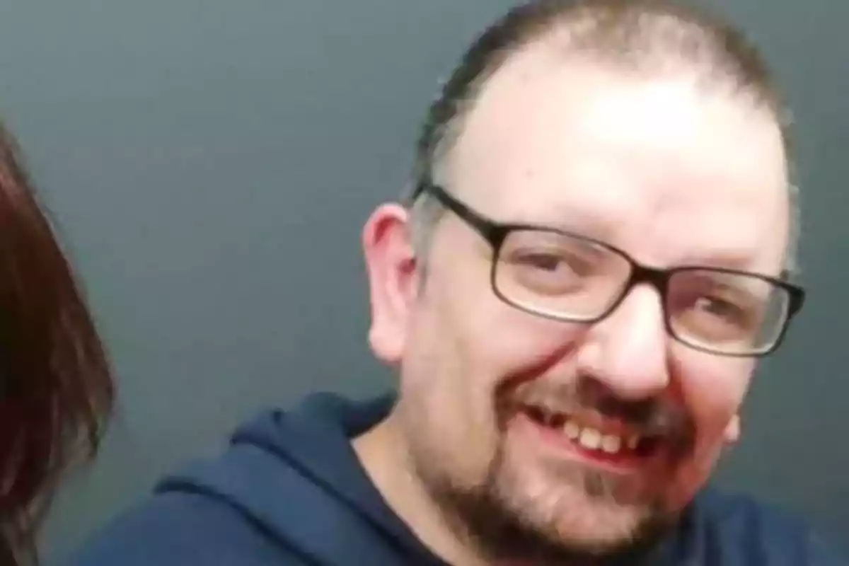 Un hombre con gafas y barba sonríe frente a un fondo gris.