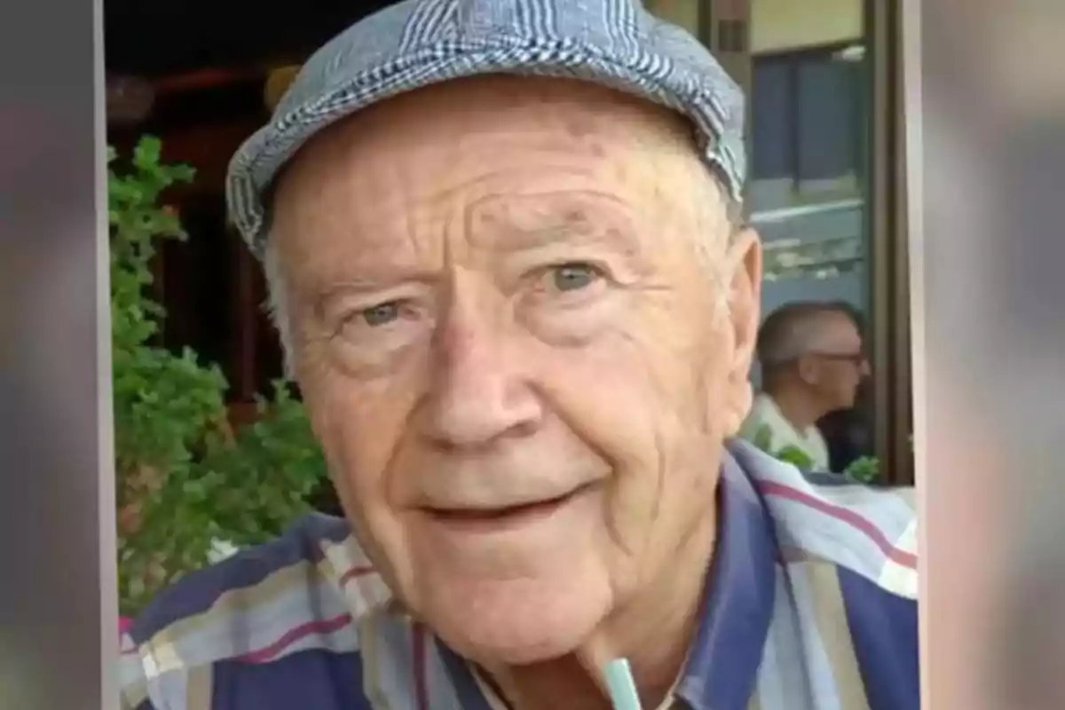 Un hombre mayor con una gorra a cuadros y una camisa de rayas, sonriendo mientras sostiene una pajilla.