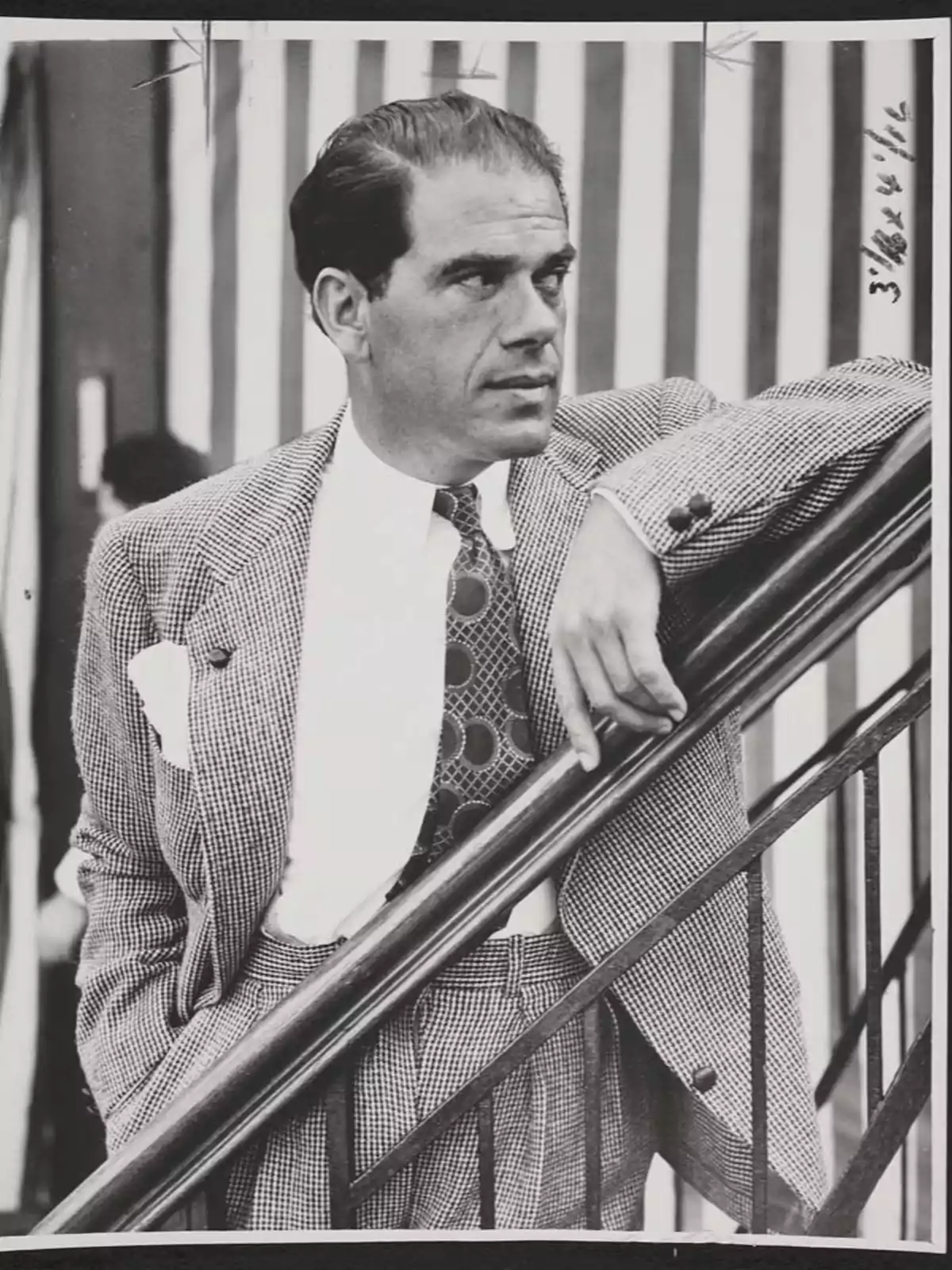 Un hombre con traje a cuadros y corbata de lunares se apoya en una barandilla mientras mira hacia un lado. Un hombre con traje a cuadros y corbata de lunares se apoya en una barandilla mientras mira hacia un lado.