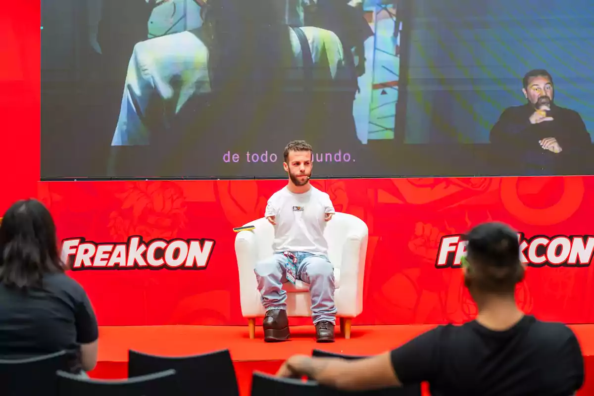 Una persona está sentada en un escenario durante un evento llamado FreakCon, con una pantalla grande detrás mostrando subtítulos y un intérprete de lenguaje de señas.