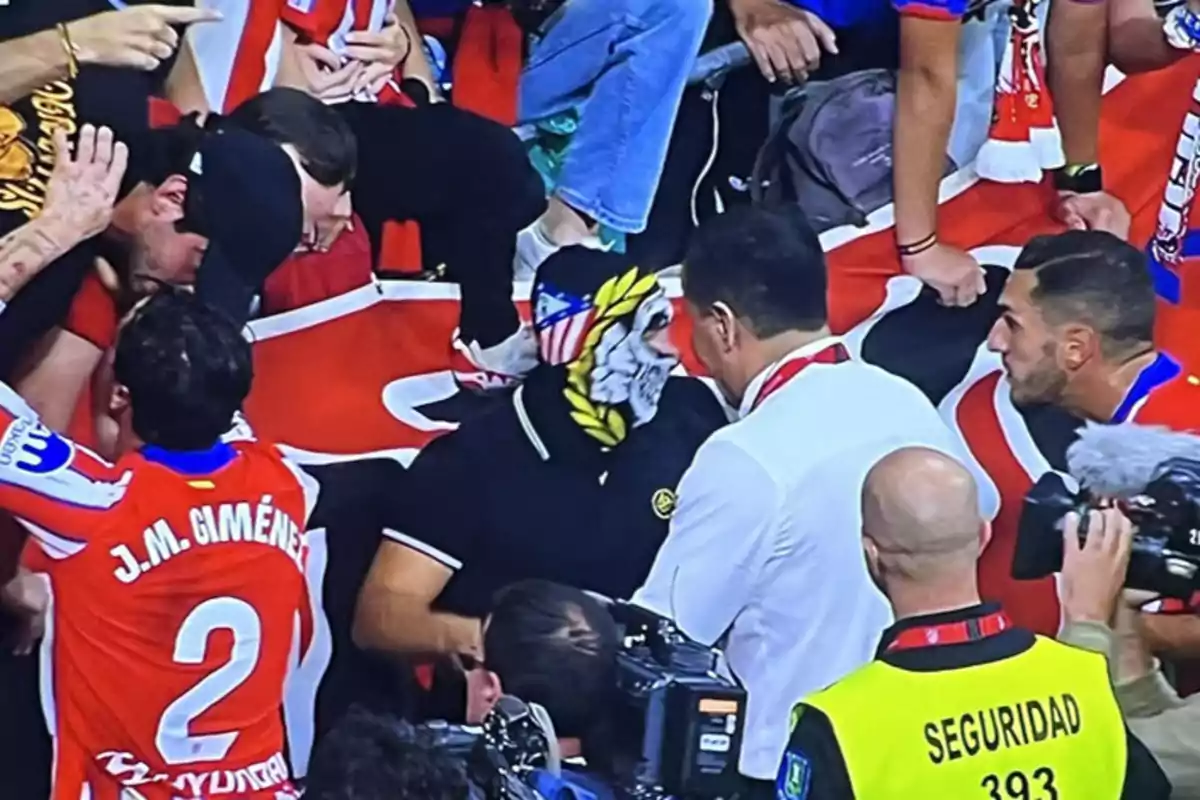 Un grupo de personas rodea a un hombre con una máscara en un evento deportivo, mientras un jugador con el número 2 en su camiseta se acerca y un miembro de seguridad observa.