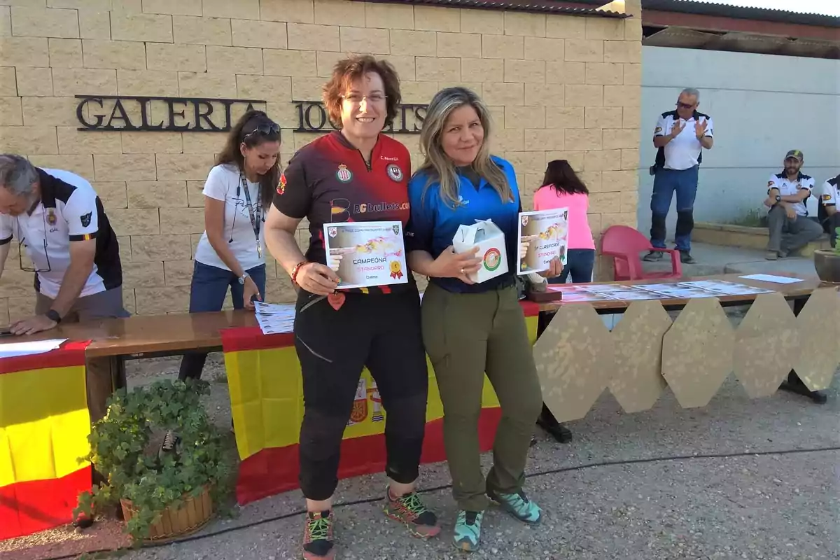 Dos mujeres posan con certificados y trofeos en un evento deportivo, mientras otras personas trabajan en una mesa detrás de ellas.