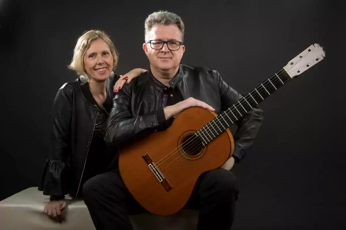 Una mujer y un hombre posan juntos, el hombre sostiene una guitarra clásica y ambos visten chaquetas de cuero negro.