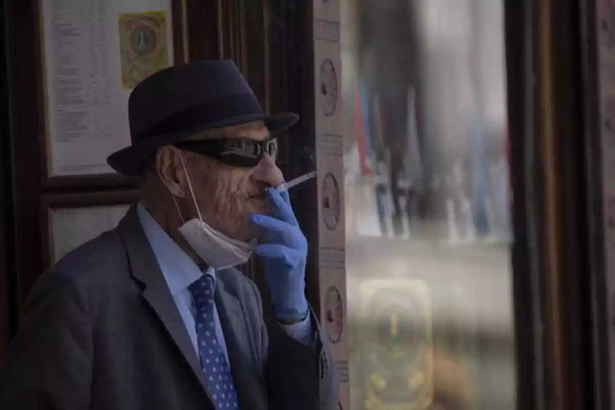 Hombre mayor con sombrero y gafas de sol fuma un cigarro mientras usa guantes azules y lleva la mascarilla bajada Hombre mayor con sombrero y gafas de sol fuma un cigarro mientras usa guantes azules y lleva la mascarilla bajada