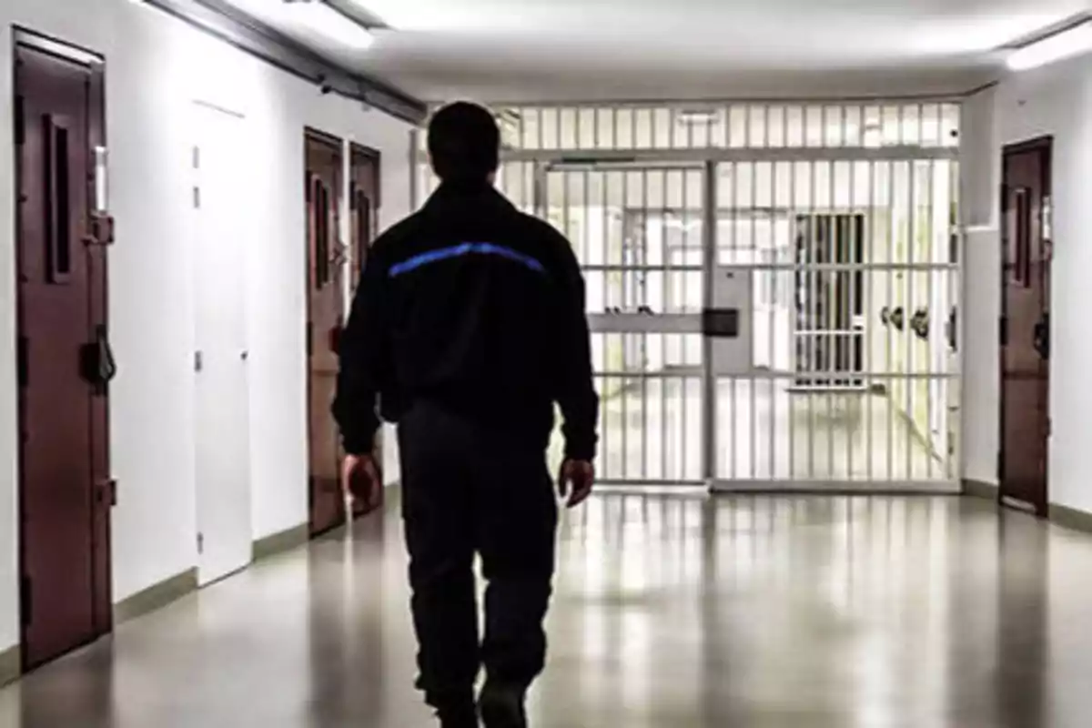 Un guardia de seguridad caminando por un pasillo de una prisión con celdas a los lados y una puerta de rejas al fondo.