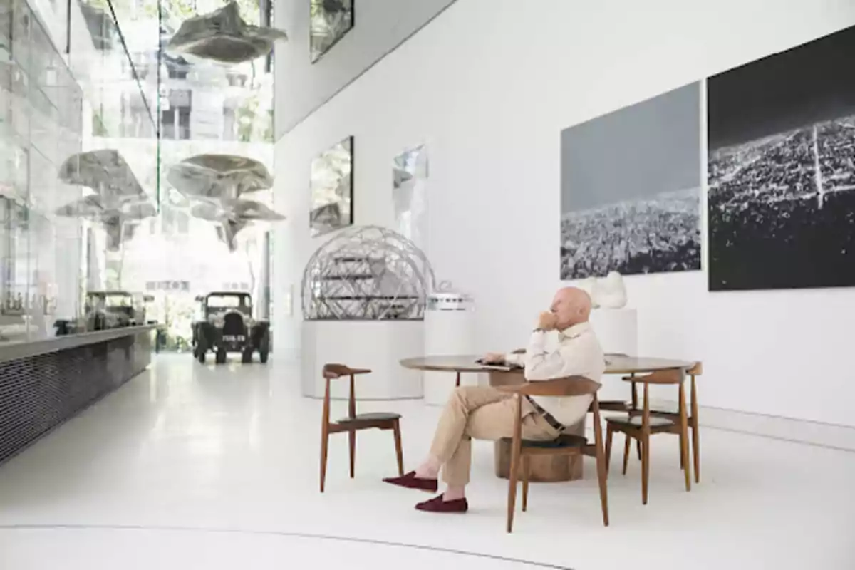 Hombre sentado en una mesa de madera en un espacio moderno y luminoso con techos altos esculturas colgantes y autos antiguos al fondo Hombre sentado en una mesa de madera en un espacio moderno y luminoso con techos altos esculturas colgantes y autos antiguos al fondo