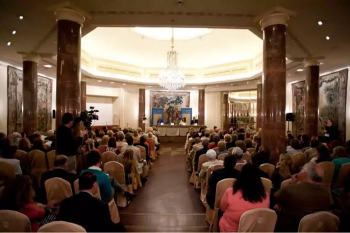 Una conferencia en un salón elegante con columnas de mármol y un candelabro, donde muchas personas están sentadas escuchando a un panel de oradores.