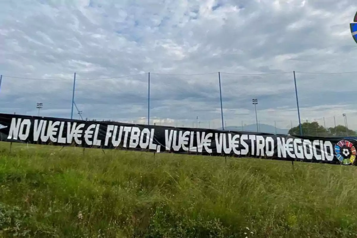 Una pancarta negra con letras blancas que dice "No vuelve el fútbol, vuelve vuestro negocio" está colocada en un campo de césped con un cielo nublado de fondo.