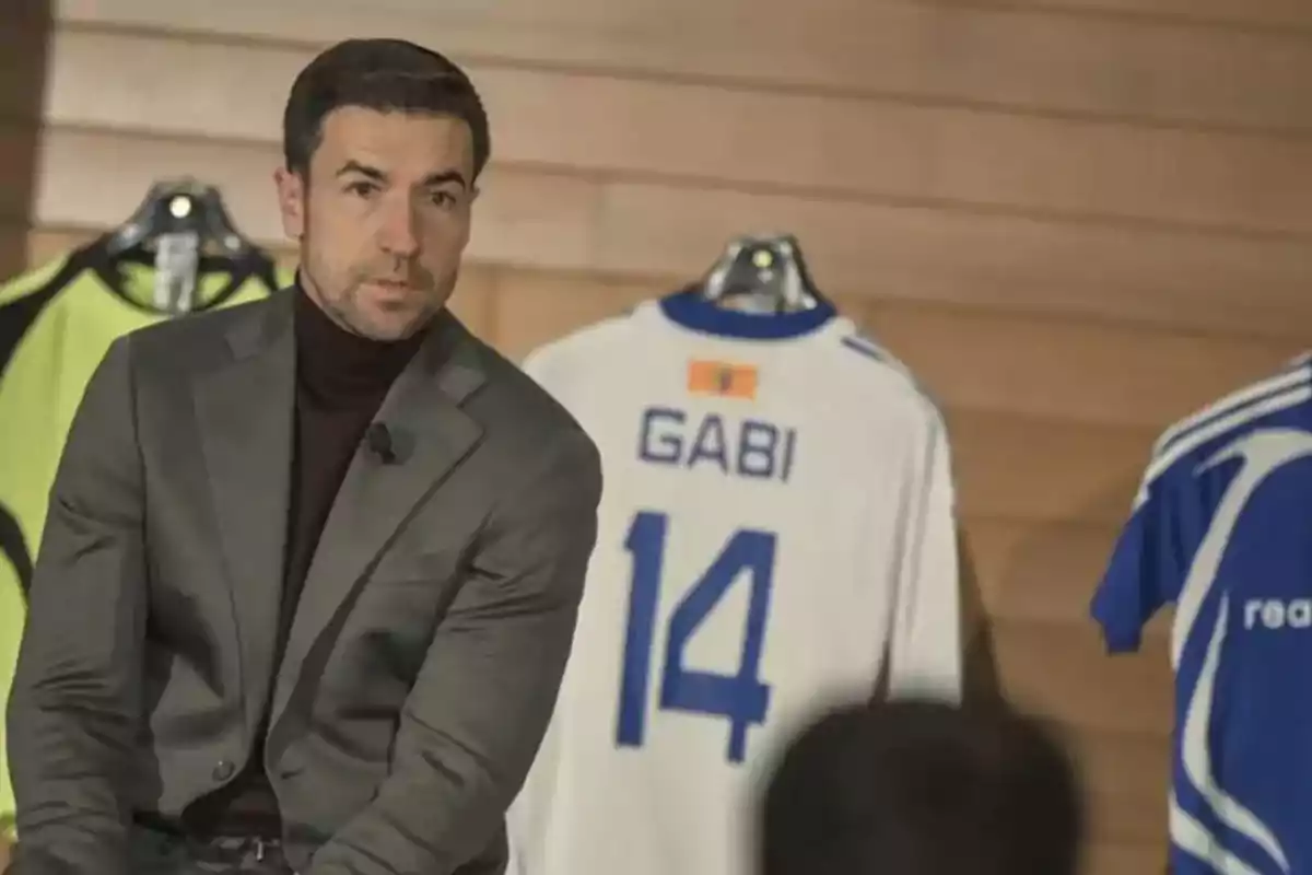 Un hombre con traje gris está sentado frente a una pared de madera, detrás de él hay una camiseta de fútbol blanca con el nombre "Gabi" y el número 14.