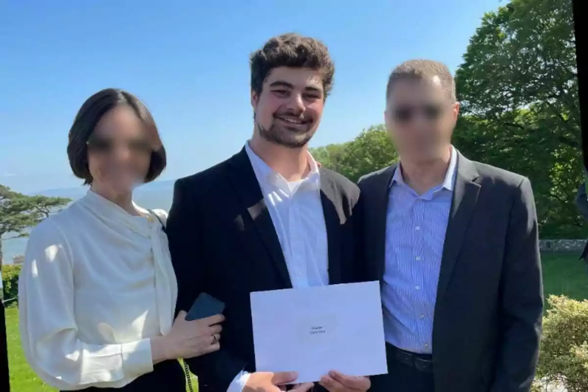 Gabriel Giacomelli sonriente sostiene un sobre mientras está de pie entre dos personas con el fondo de un paisaje verde y cielo azul.