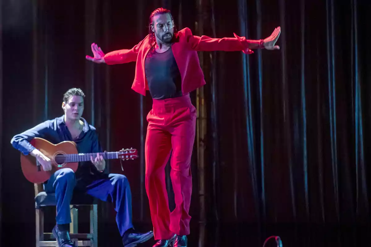 Un bailarín de flamenco vestido de rojo realiza un movimiento con los brazos extendidos mientras un guitarrista toca en el fondo.