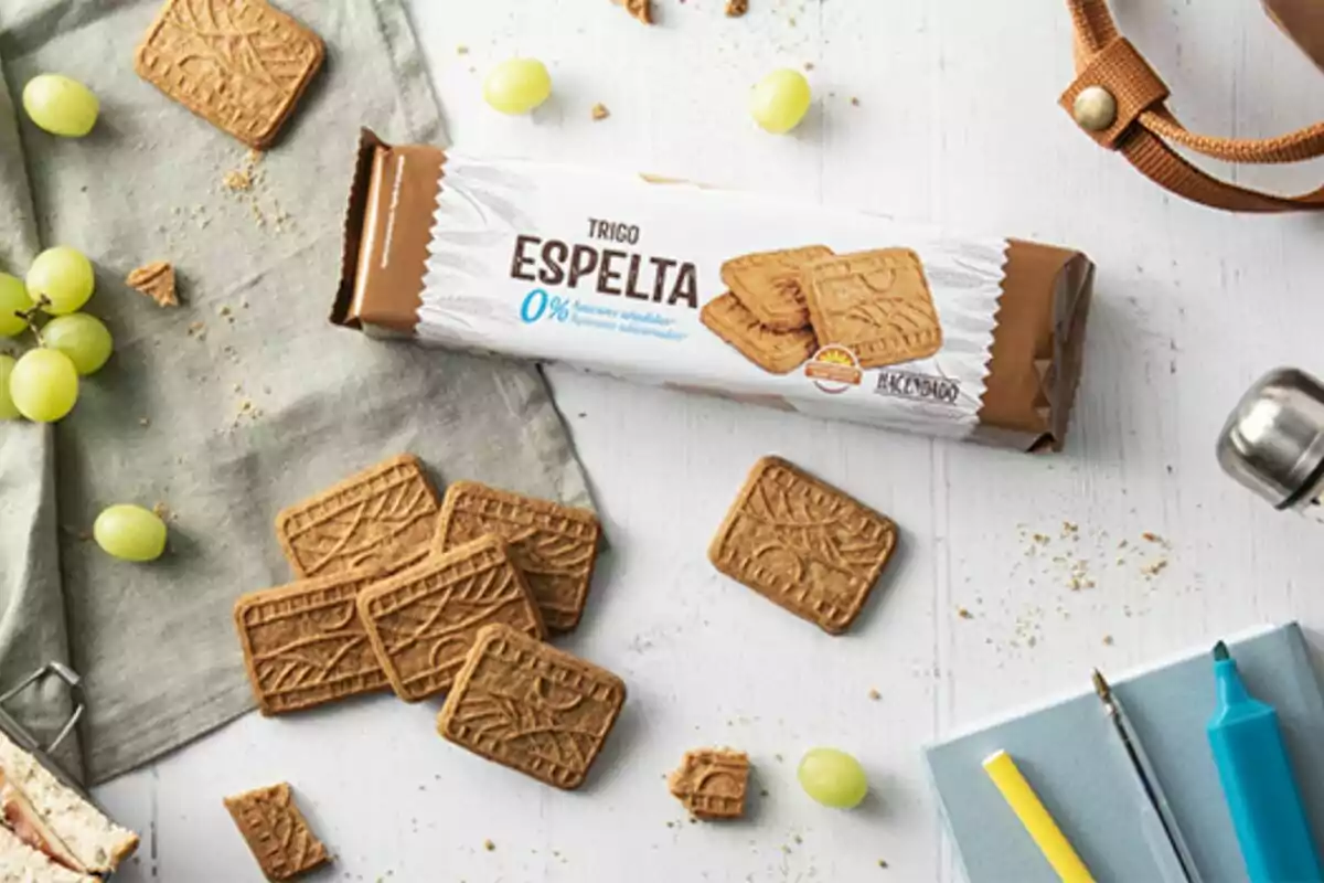 Paquete de galletas de trigo espelta de Mercadona sobre una mesa blanca con algunas galletas sueltas, uvas verdes y artículos de papelería alrededor. Paquete de galletas de trigo espelta de Mercadona sobre una mesa blanca con algunas galletas sueltas, uvas verdes y artículos de papelería alrededor.