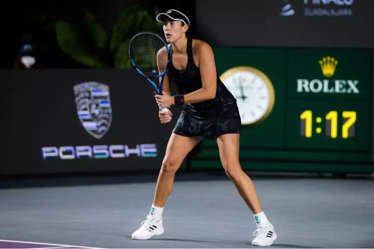 Jugadora de tenis en posición de espera durante un partido, con una raqueta en las manos y vestida con ropa deportiva negra, en una cancha con logotipos de Porsche y Rolex en el fondo.