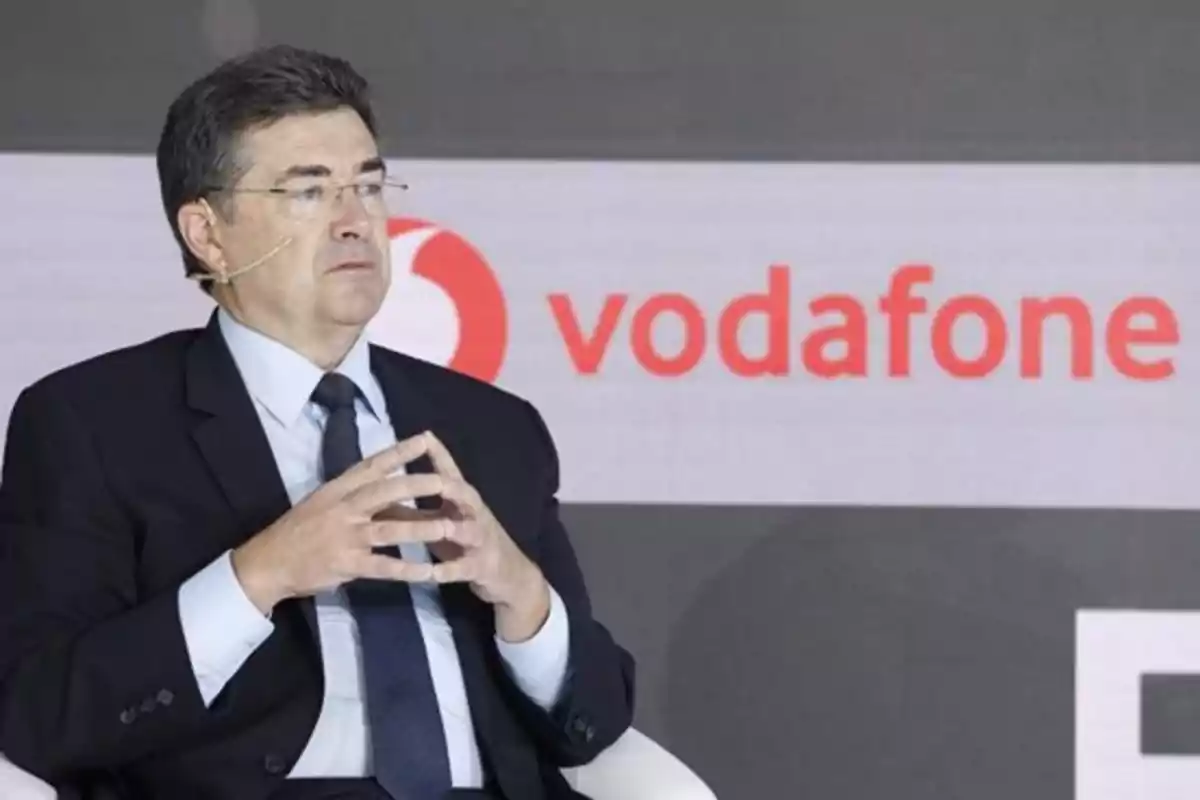 Man in a suit with glasses and a headset microphone sitting in front of a background with the Vodafone logo Man in a suit with glasses and a headset microphone sitting in front of a background with the Vodafone logo