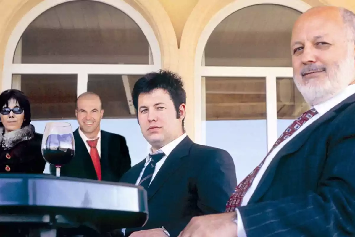 Un grupo de personas vestidas formalmente posando frente a un edificio con ventanas arqueadas y una copa de vino en primer plano.