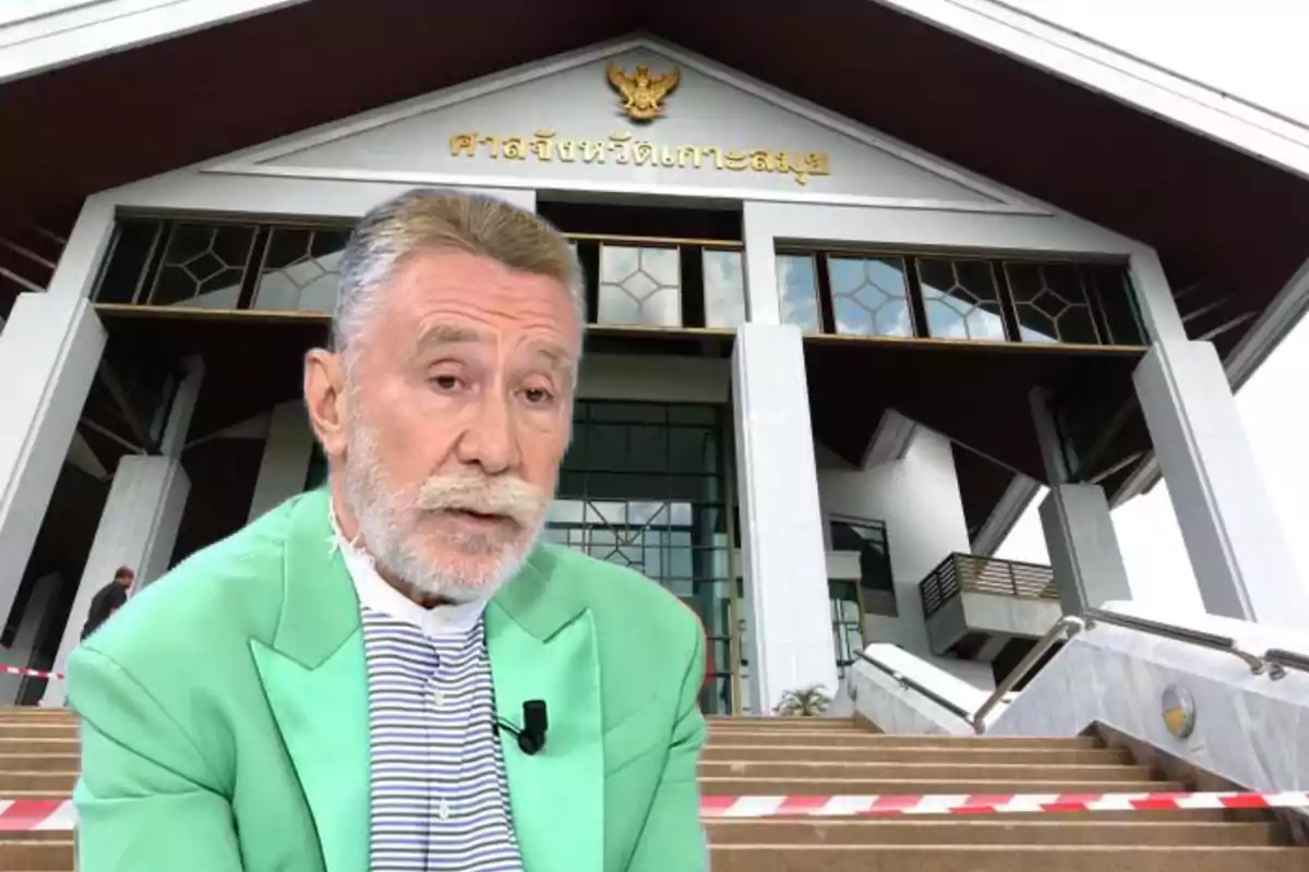 Un hombre con barba y bigote, vestido con una chaqueta verde y una camisa a rayas, está frente a un edificio con arquitectura moderna y un letrero en un idioma extranjero.