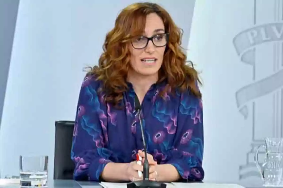Mujer de cabello rizado y gafas hablando frente a un micrófono en una mesa de conferencia