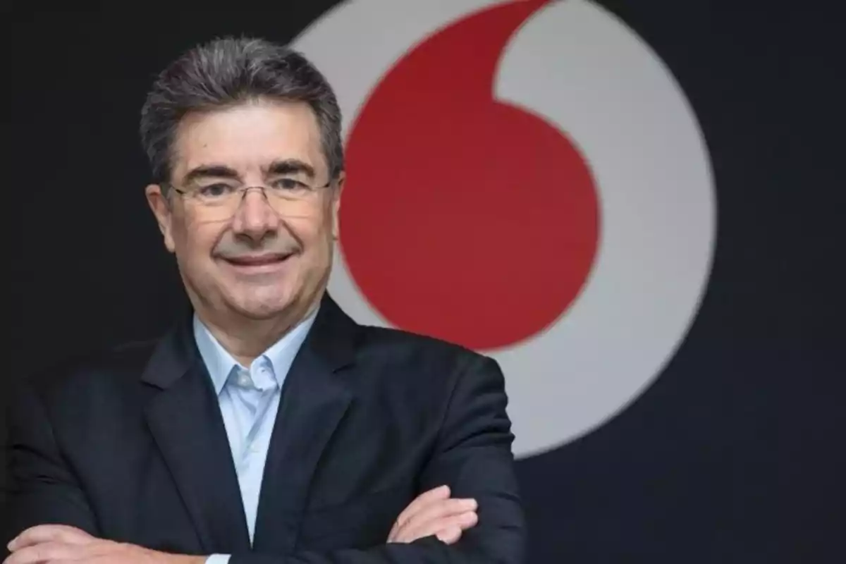 Hombre con gafas y traje oscuro sonriendo frente a un logotipo circular rojo y blanco.