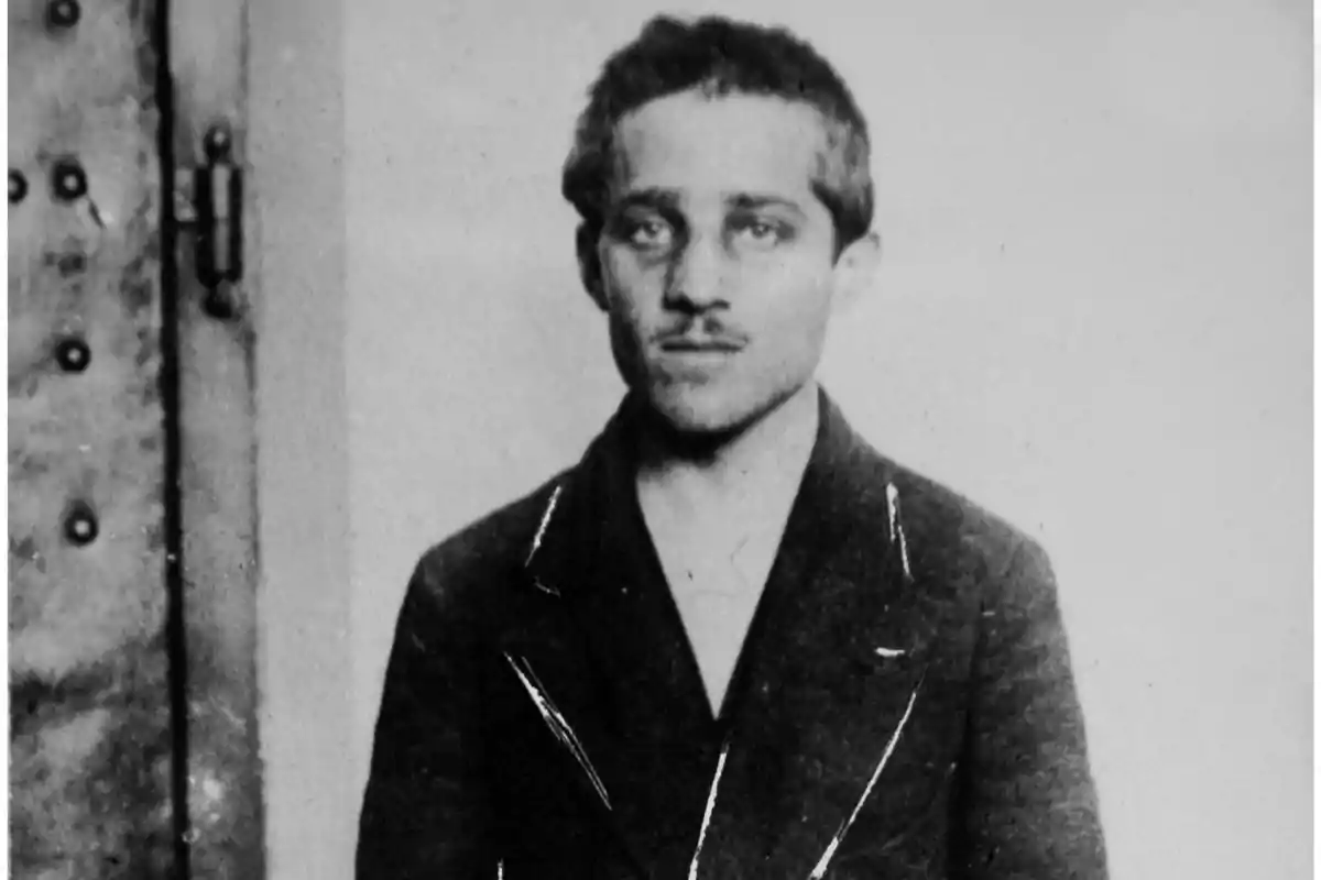 Un hombre joven con bigote y cabello corto viste un traje oscuro y mira hacia la cámara en una fotografía en blanco y negro.