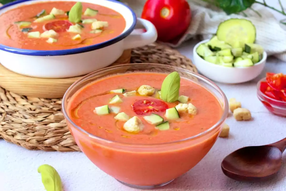 Un delicioso gazpacho servido en un tazón de vidrio, adornado con trozos de pepino, tomate, crutones y una hoja de albahaca, acompañado de un tazón adicional de gazpacho y vegetales frescos en el fondo.