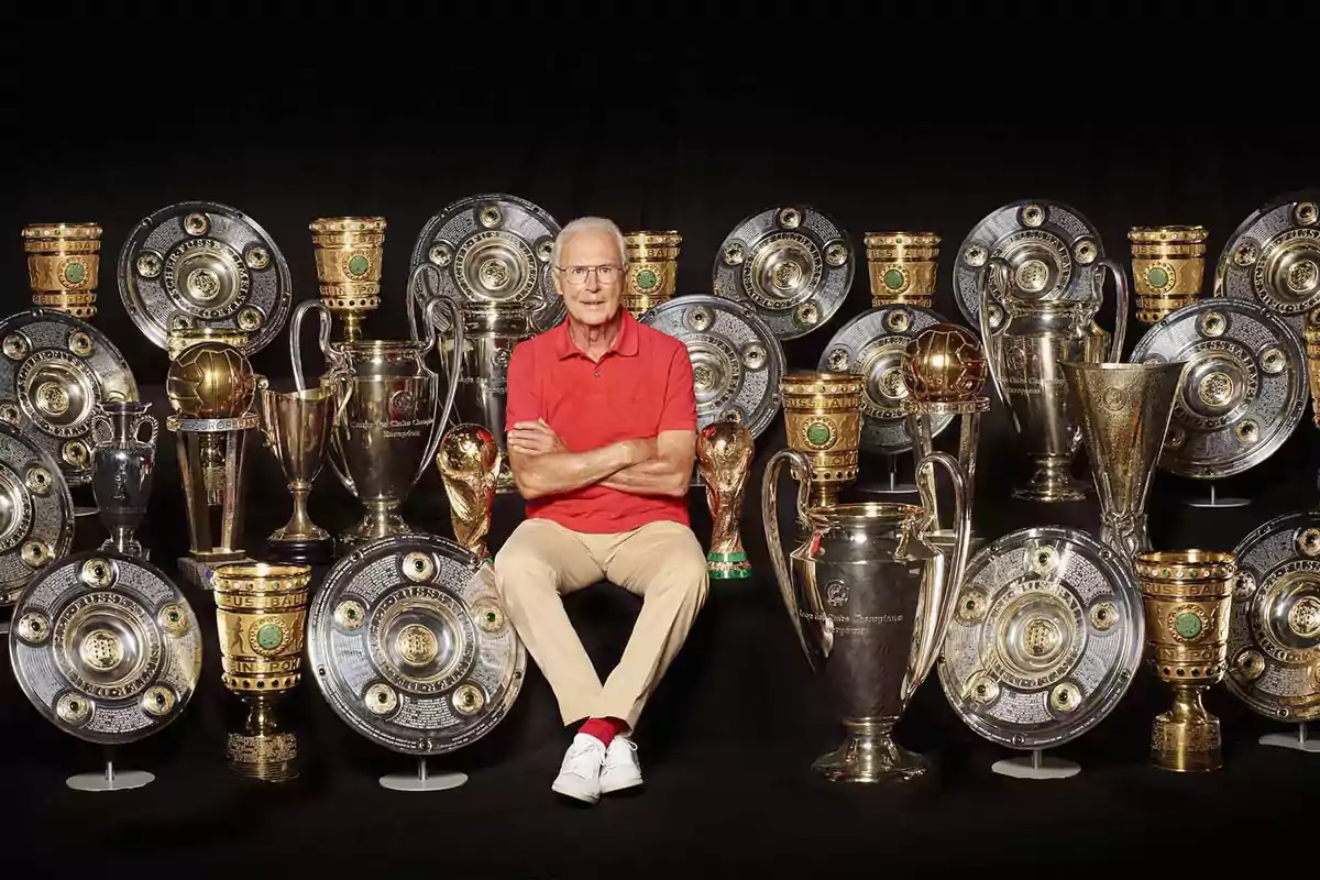 Un hombre mayor con camisa roja y pantalones beige está sentado con los brazos cruzados, rodeado de numerosos trofeos y copas de fútbol.