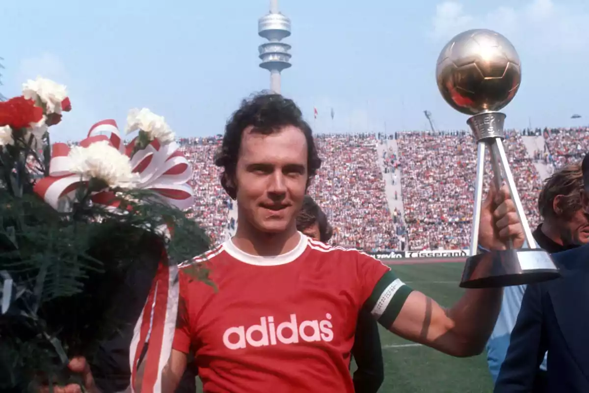 Un jugador de fútbol con una camiseta roja de Adidas sostiene un trofeo y un ramo de flores en un estadio lleno de espectadores.