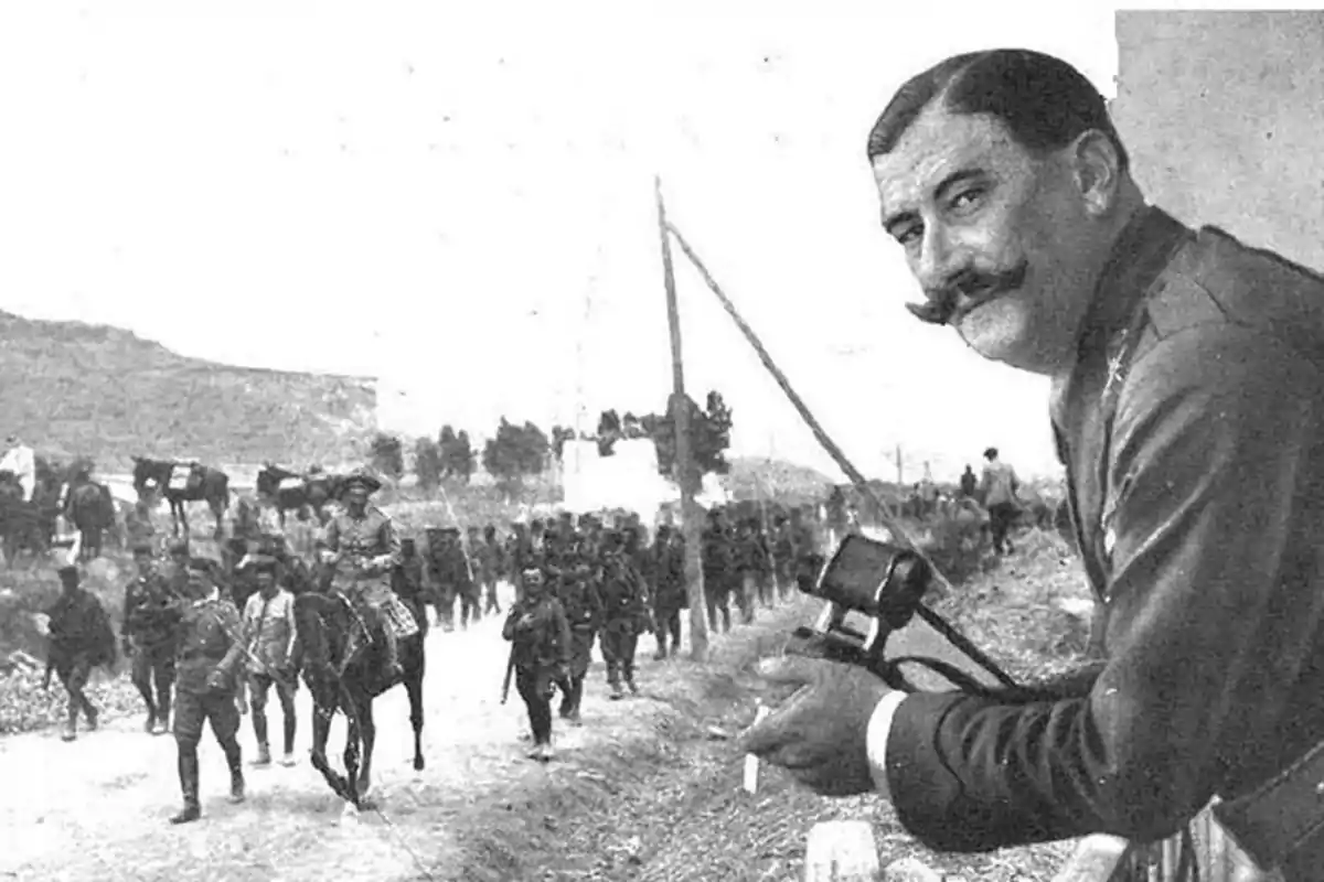 Un hombre con bigote y uniforme militar observa a un grupo de soldados marchando por un camino mientras sostiene un par de binoculares.