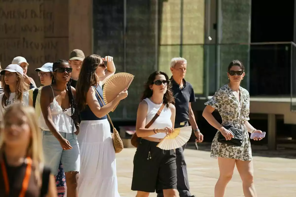 Grupo de personas caminando bajo el sol, algunas usando abanicos y gafas de sol para protegerse del calor. Grupo de personas caminando bajo el sol, algunas usando abanicos y gafas de sol para protegerse del calor.