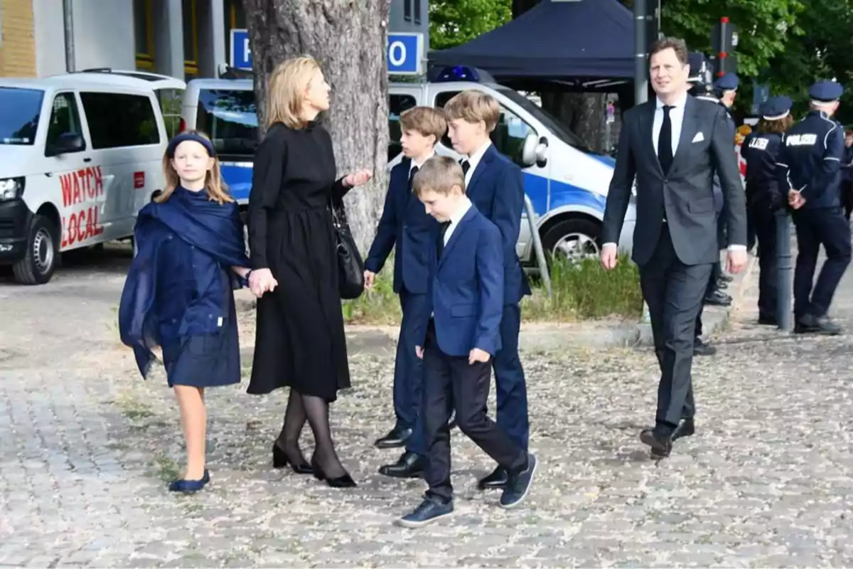 Un grupo de personas vestidas formalmente camina por una calle empedrada con vehículos y policías al fondo. Un grupo de personas vestidas formalmente camina por una calle empedrada con vehículos y policías al fondo.